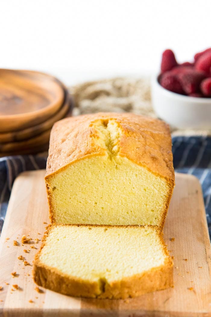 A sliced classic pound cake on a wooden board, with serving plates and a bowl of raspberries in the background. 