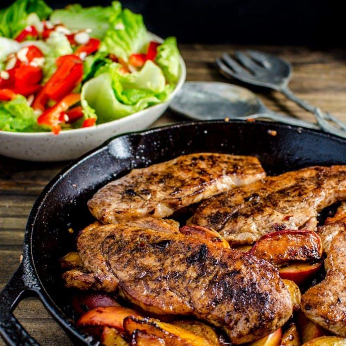 Pan fried pork chops with peach halves in a black cast iron pan with a salad in the background.