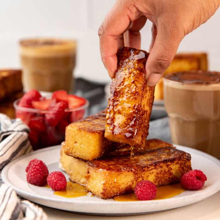 A hand picking up a French toast stick that's dripping with maple syrup onto a plate with more French toast sticks.