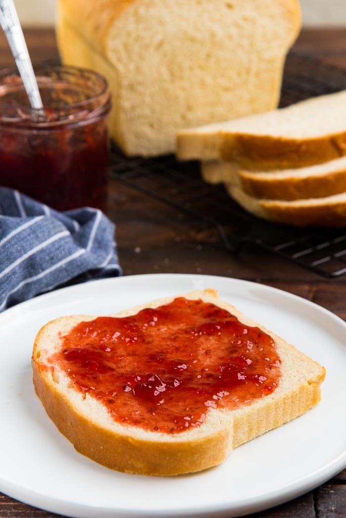 A slice of homemade bread on a white plate with jam spread on top, with the rest of the bread loaf in the background.