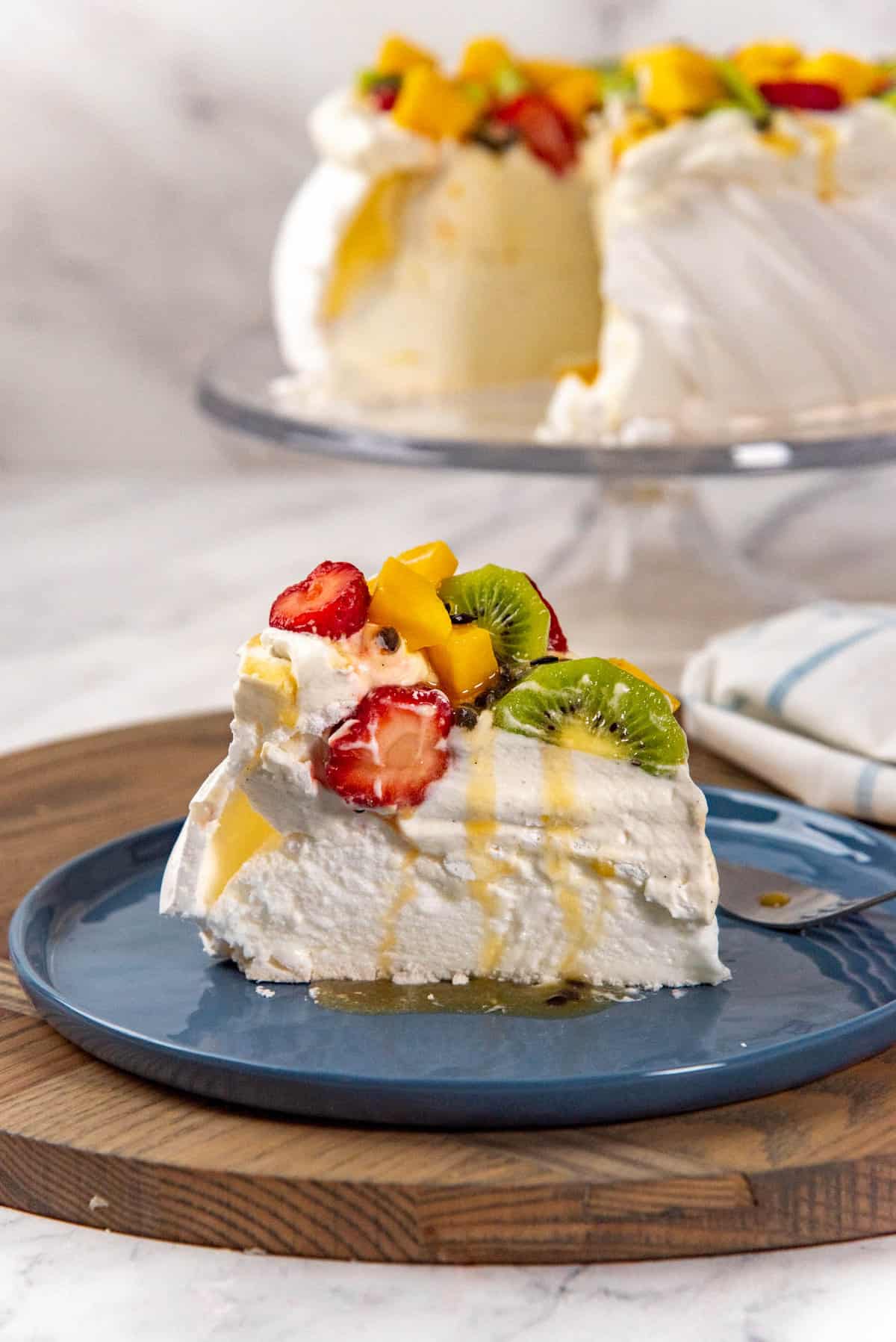 A slice of pavlova served on a blue plate with the bigger pavlova on the cake stand in the background. The slice of pavlova shows the marshmallow center and the crisp meringue outer shell as well as the fresh fruits and whipped cream on top.